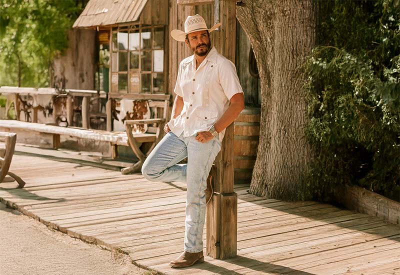 A man wearing the Antonio 13" Western boot in brown leaning against a fence.